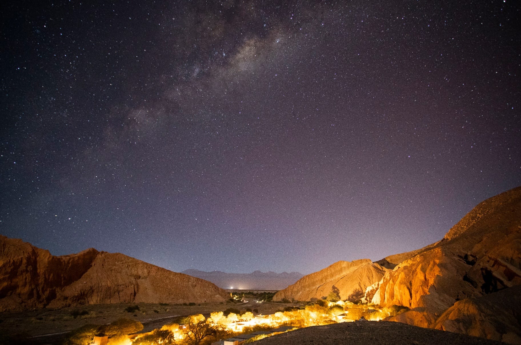 Via Láctea sobre oásis iluminado entre encostas no deserto noturno